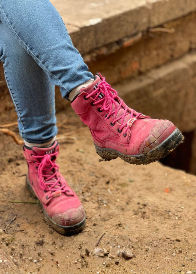 Person sitting on worksite in pink lace-up composite work boots, covered in mud.