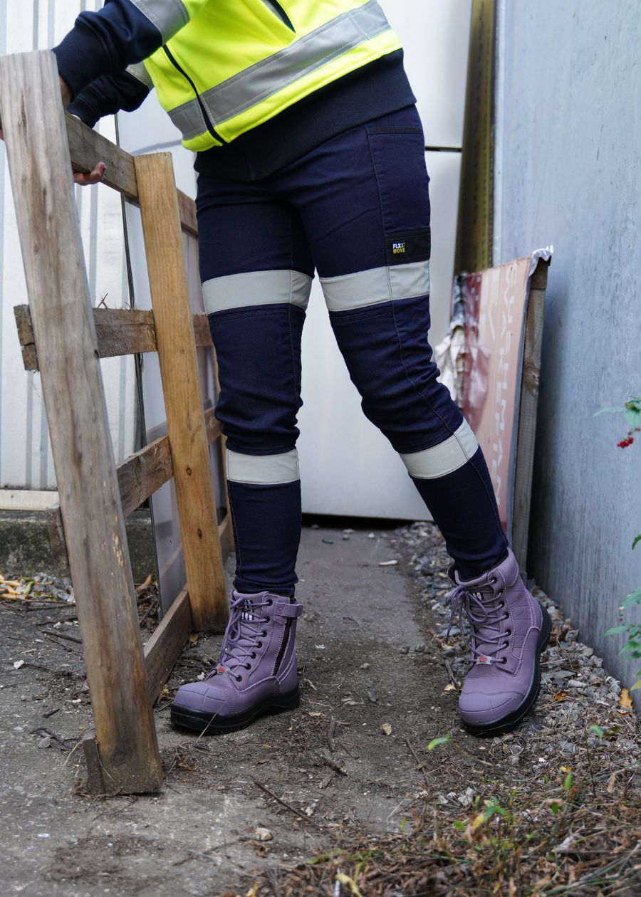 A woman in workwear is carrying a wooden palette while wearing purple zip-up safety boots.  