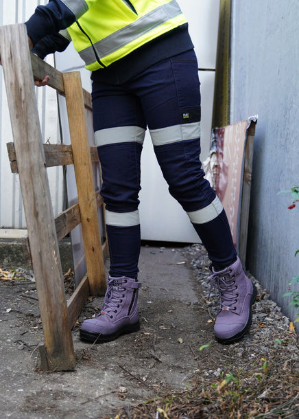 A woman in workwear is carrying a wooden palette while wearing purple zip-up safety boots.  