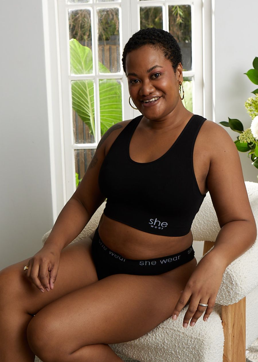 Smiling woman seated by a window, wearing a matching set of stretchy black organic bamboo underwear and crop top. She looks relaxed and comfortable.
