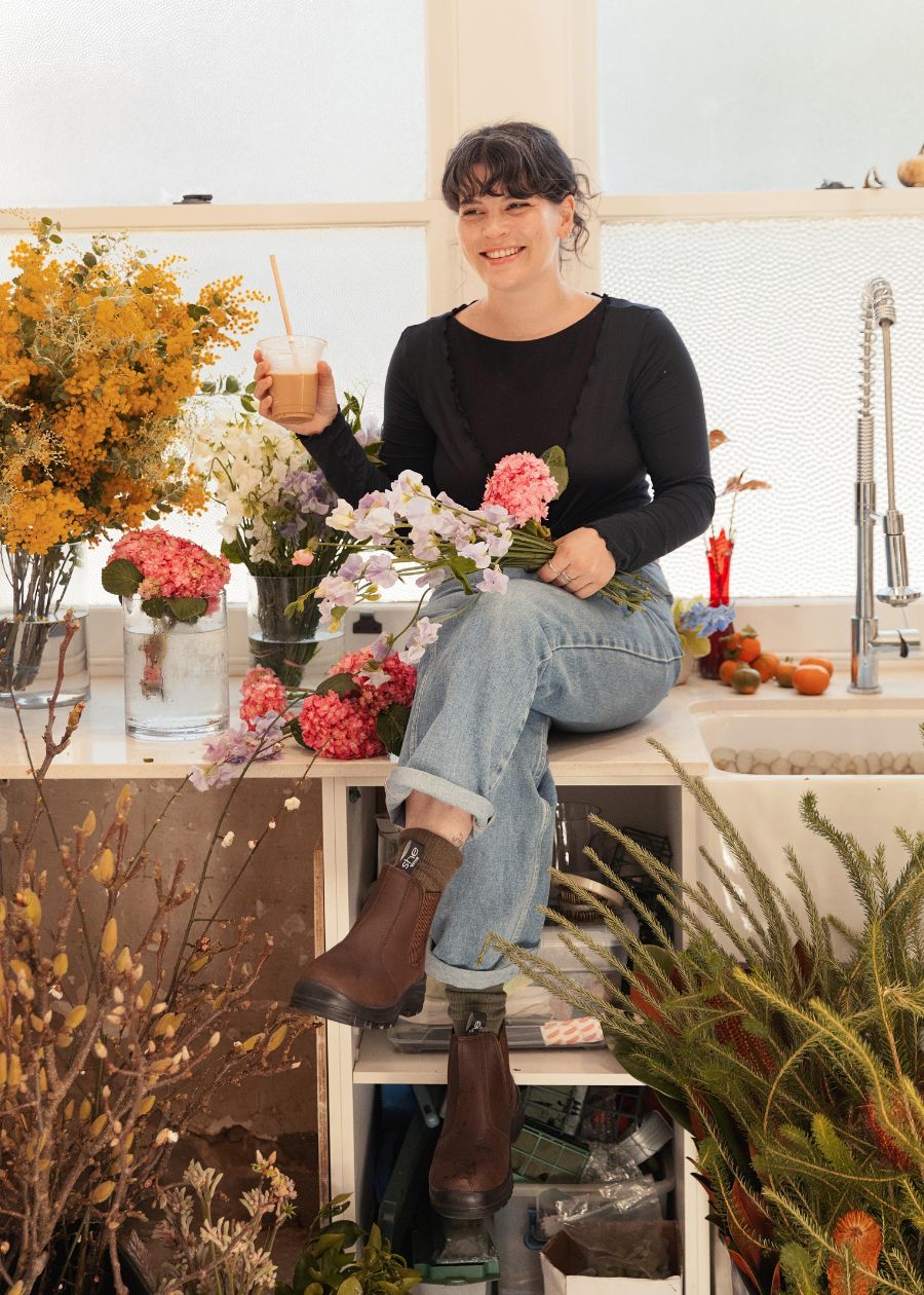Woman sitting down in a florist shop with brown working boots on holding flowers