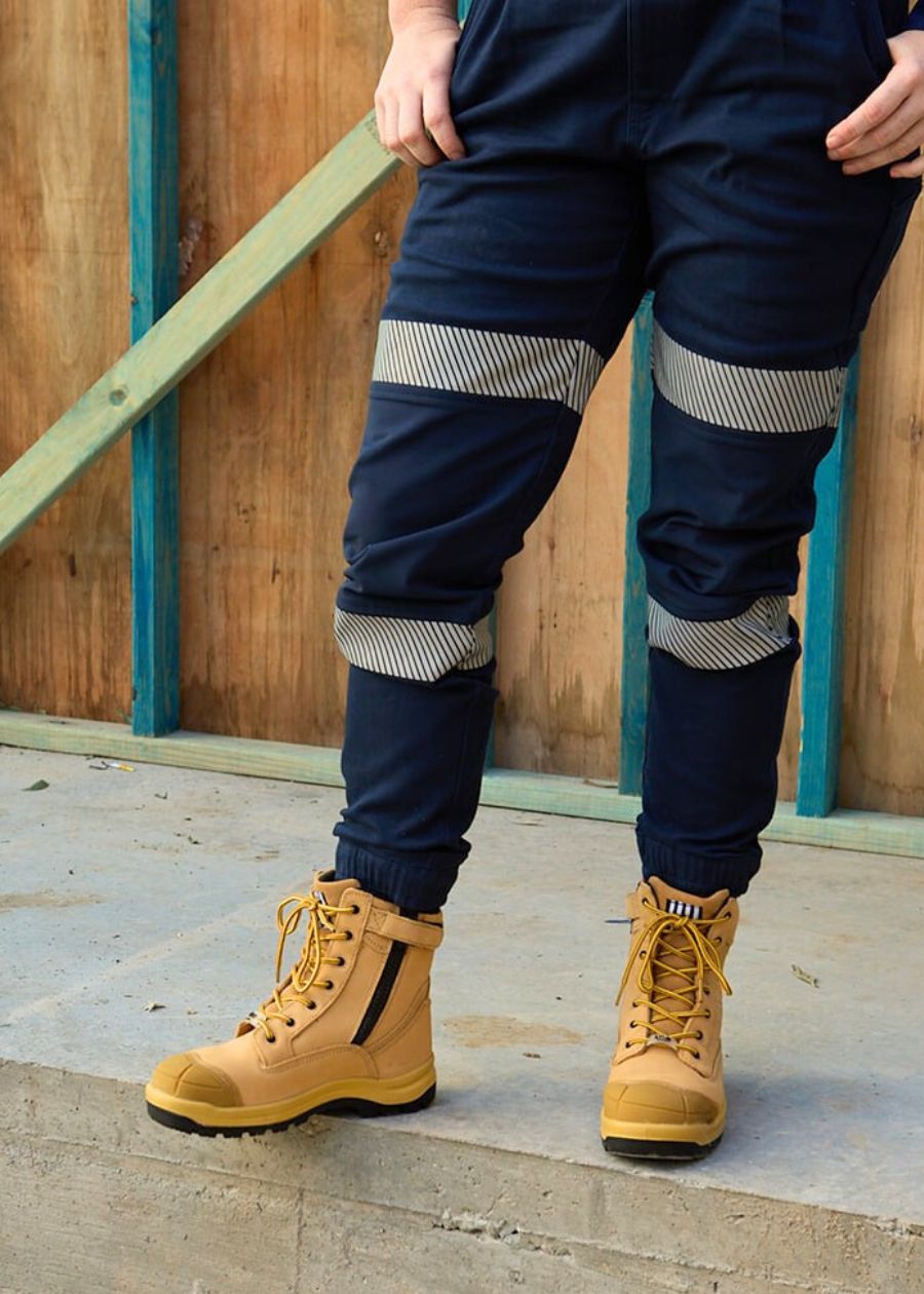 Person wearing tan lace-up work boots with black soles and navy pants featuring reflective stripes, standing on a concrete surface in front of a wooden frame.