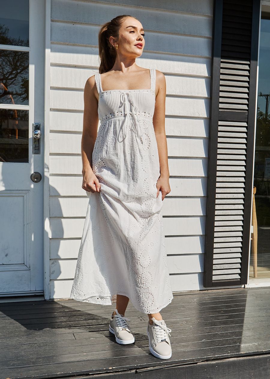 Woman in white dress with beige lace-up sneakers. Sun in face, with house in background.